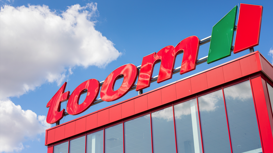 Außenaufnahme eines toom-Baumarkts aus niedriger Perspektive. Über der rot verkleideten Glasfassade steht der große rote „toom“-Schriftzug mit grün-rotem Logo vor blauem Himmel und weißen Wolken.