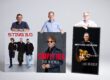 Drei Männer stehen vor einer hellgrauen Wand und präsentieren große Konzertplakate. Diese kündigen Auftritte von Sting 3.0, Simply Red und Bryan Adams in Hemer an. Die Männer lächeln in die Kamera und stützen ihre Hände auf die Plakate.