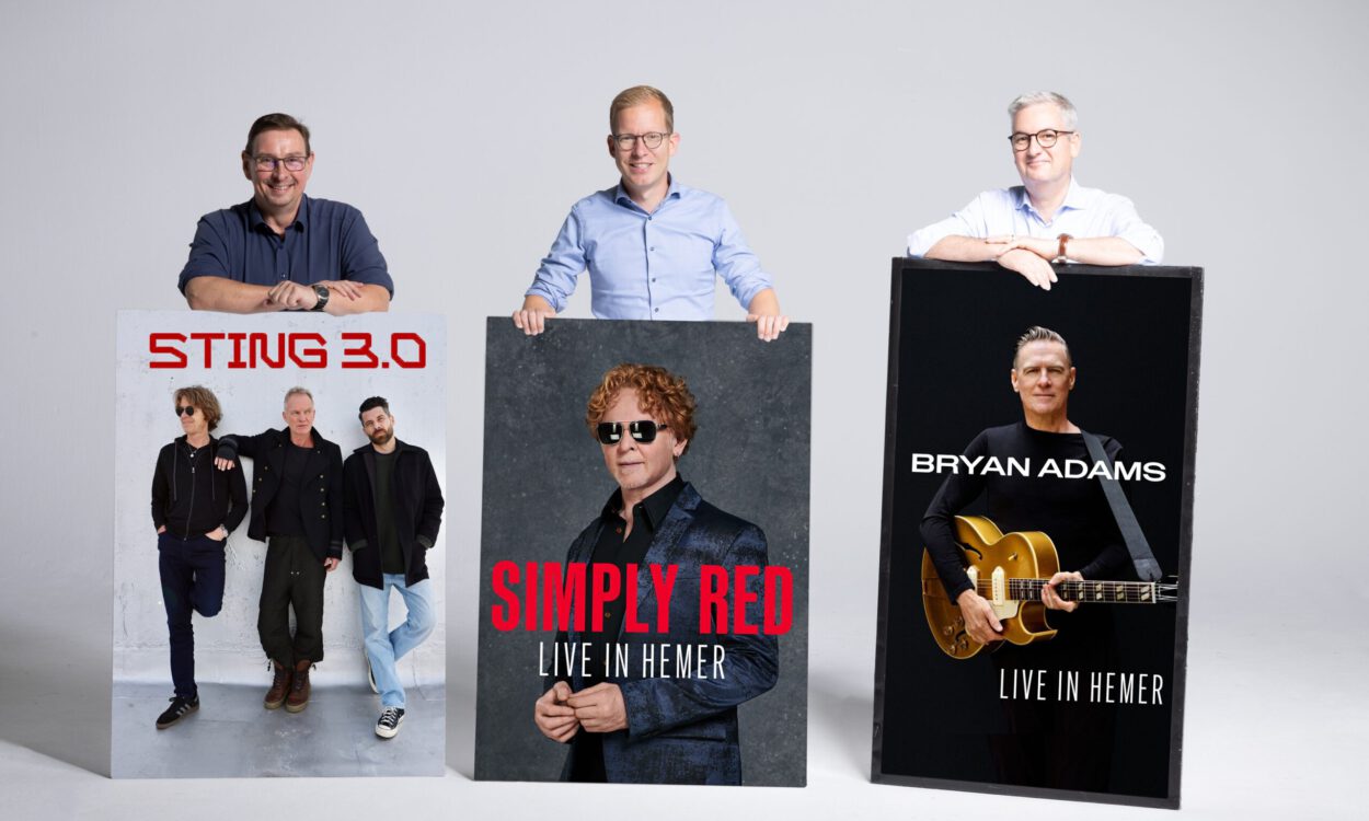 Drei Männer stehen vor einer hellgrauen Wand und präsentieren große Konzertplakate. Diese kündigen Auftritte von Sting 3.0, Simply Red und Bryan Adams in Hemer an. Die Männer lächeln in die Kamera und stützen ihre Hände auf die Plakate.