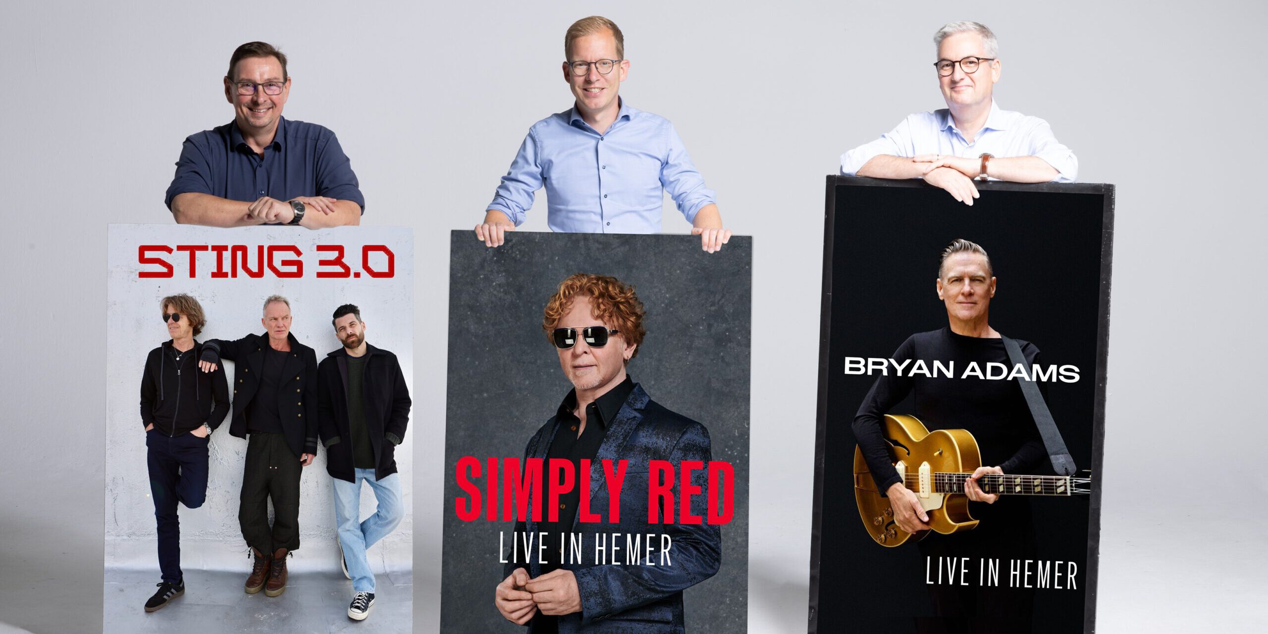 Drei Männer stehen vor einer hellgrauen Wand und präsentieren große Konzertplakate. Diese kündigen Auftritte von Sting 3.0, Simply Red und Bryan Adams in Hemer an. Die Männer lächeln in die Kamera und stützen ihre Hände auf die Plakate.