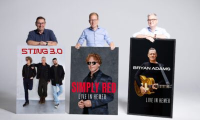 Drei Männer stehen vor einer hellgrauen Wand und präsentieren große Konzertplakate. Diese kündigen Auftritte von Sting 3.0, Simply Red und Bryan Adams in Hemer an. Die Männer lächeln in die Kamera und stützen ihre Hände auf die Plakate.