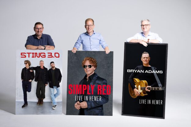 Drei Männer stehen vor einer hellgrauen Wand und präsentieren große Konzertplakate. Diese kündigen Auftritte von Sting 3.0, Simply Red und Bryan Adams in Hemer an. Die Männer lächeln in die Kamera und stützen ihre Hände auf die Plakate.