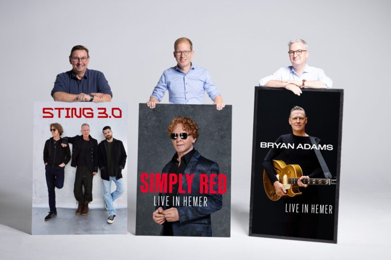 Foto: Tölle Studios Drei Männer stehen vor einer hellgrauen Wand und präsentieren große Konzertplakate. Diese kündigen Auftritte von Sting 3.0, Simply Red und Bryan Adams in Hemer an. Die Männer lächeln in die Kamera und stützen ihre Hände auf die Plakate.