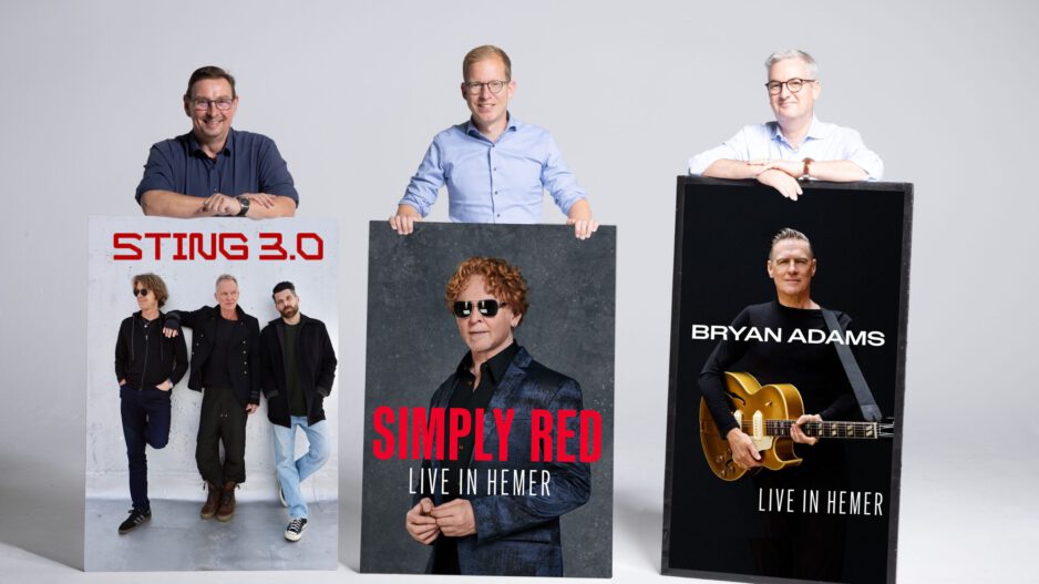 Drei Männer stehen vor einer hellgrauen Wand und präsentieren große Konzertplakate. Diese kündigen Auftritte von Sting 3.0, Simply Red und Bryan Adams in Hemer an. Die Männer lächeln in die Kamera und stützen ihre Hände auf die Plakate.