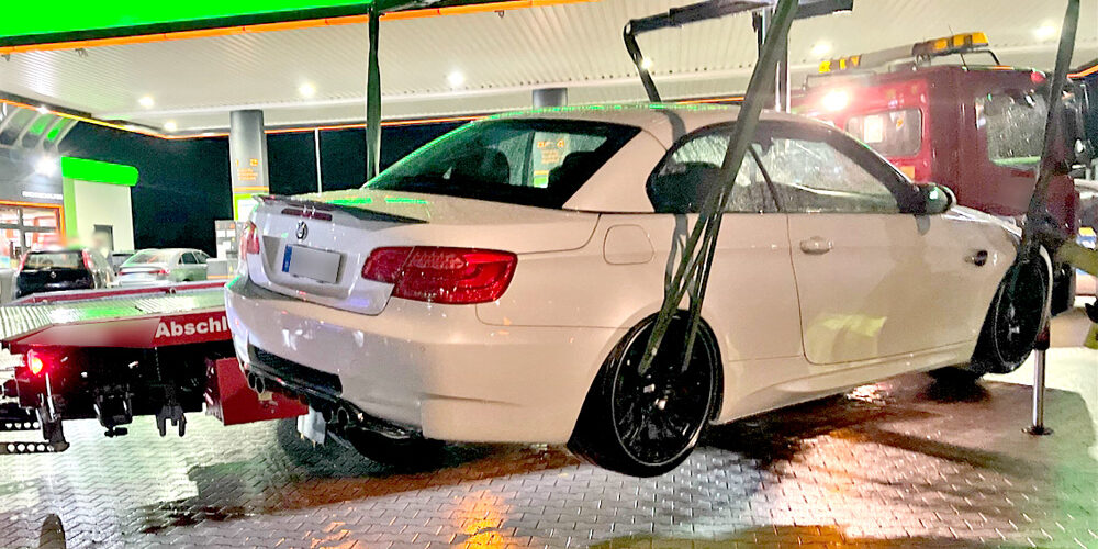 Eine weiße BMW-Limousine mit schwarzen Felgen wird nachts an einer hell beleuchteten Tankstelle von einem Abschleppwagen angehoben. Im Hintergrund steht ein rotes Abschleppfahrzeug. Der Boden ist nass und spiegelt die Lichter der Tankstelle.