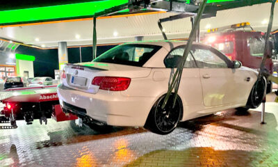 Eine weiße BMW-Limousine mit schwarzen Felgen wird nachts an einer hell beleuchteten Tankstelle von einem Abschleppwagen angehoben. Im Hintergrund steht ein rotes Abschleppfahrzeug. Der Boden ist nass und spiegelt die Lichter der Tankstelle.