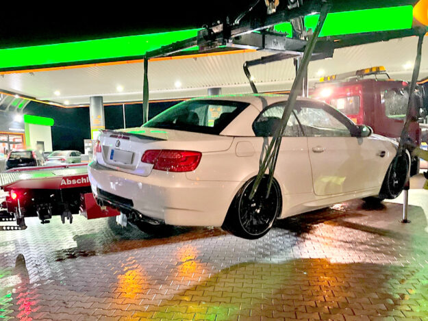 Eine weiße BMW-Limousine mit schwarzen Felgen wird nachts an einer hell beleuchteten Tankstelle von einem Abschleppwagen angehoben. Im Hintergrund steht ein rotes Abschleppfahrzeug. Der Boden ist nass und spiegelt die Lichter der Tankstelle.