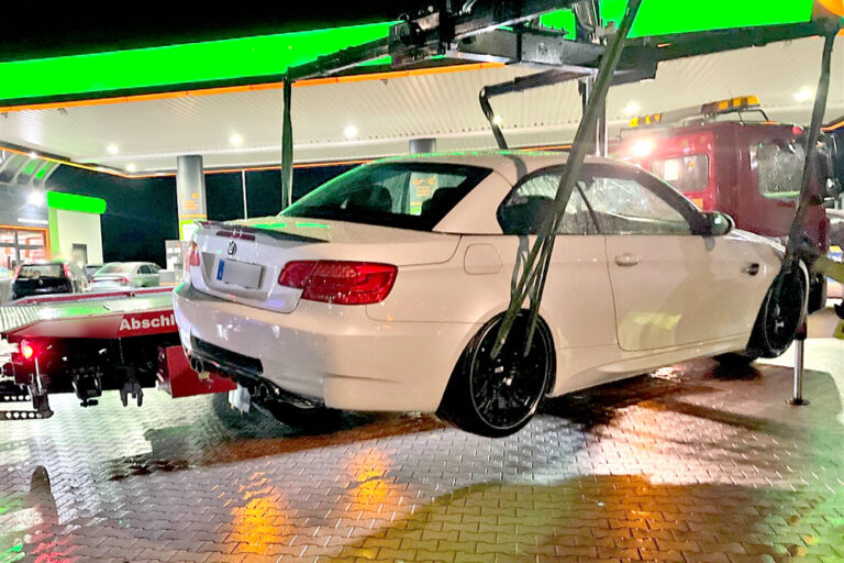 Eine weiße BMW-Limousine mit schwarzen Felgen wird nachts an einer hell beleuchteten Tankstelle von einem Abschleppwagen angehoben. Im Hintergrund steht ein rotes Abschleppfahrzeug. Der Boden ist nass und spiegelt die Lichter der Tankstelle.