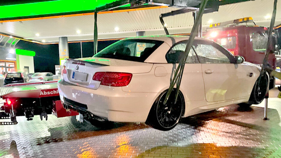 Eine weiße BMW-Limousine mit schwarzen Felgen wird nachts an einer hell beleuchteten Tankstelle von einem Abschleppwagen angehoben. Im Hintergrund steht ein rotes Abschleppfahrzeug. Der Boden ist nass und spiegelt die Lichter der Tankstelle.