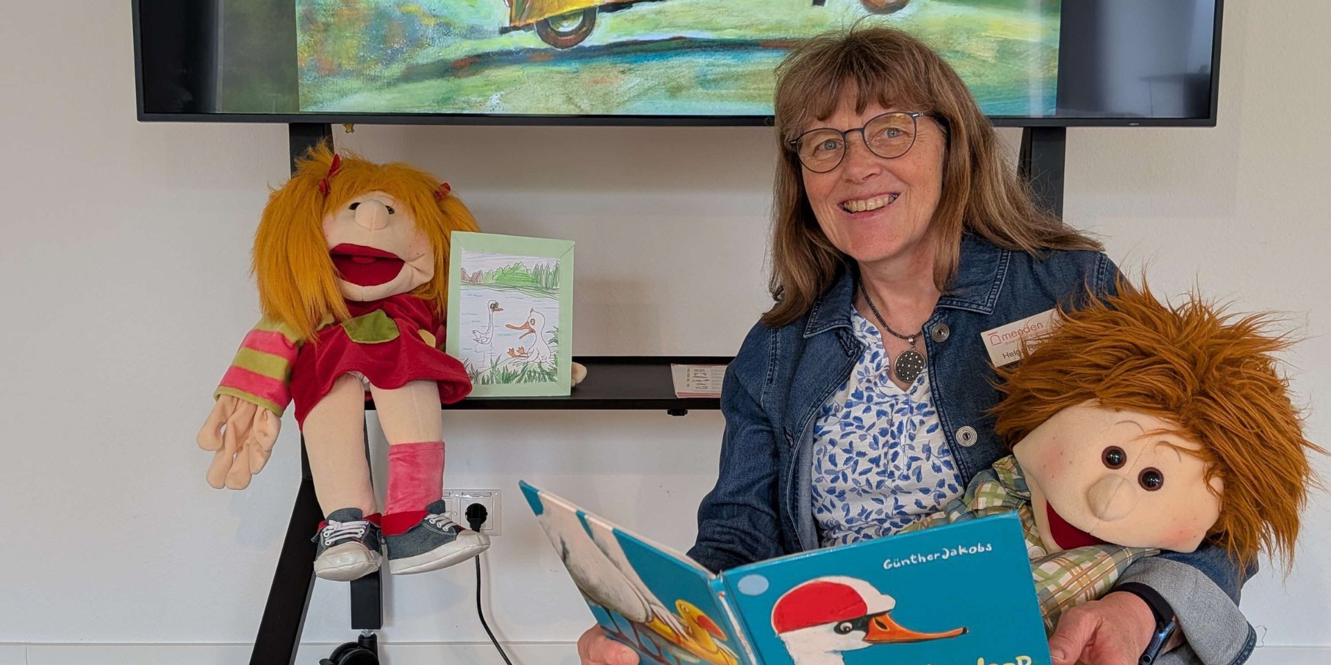 Eine lächelnde Frau sitzt mit zwei Handpuppen vor einem großen Bildschirm und hält das Bilderbuch „Schnabbeldiplapp“ geöffnet in den Händen. Auf dem Bildschirm ist eine farbenfrohe Illustration aus dem Buch mit zwei Gänsen in einem gelben Auto zu sehen.