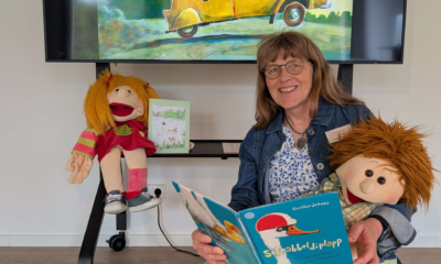 Eine lächelnde Frau sitzt mit zwei Handpuppen vor einem großen Bildschirm und hält das Bilderbuch „Schnabbeldiplapp“ geöffnet in den Händen. Auf dem Bildschirm ist eine farbenfrohe Illustration aus dem Buch mit zwei Gänsen in einem gelben Auto zu sehen.