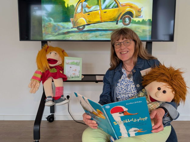 Eine lächelnde Frau sitzt mit zwei Handpuppen vor einem großen Bildschirm und hält das Bilderbuch „Schnabbeldiplapp“ geöffnet in den Händen. Auf dem Bildschirm ist eine farbenfrohe Illustration aus dem Buch mit zwei Gänsen in einem gelben Auto zu sehen.