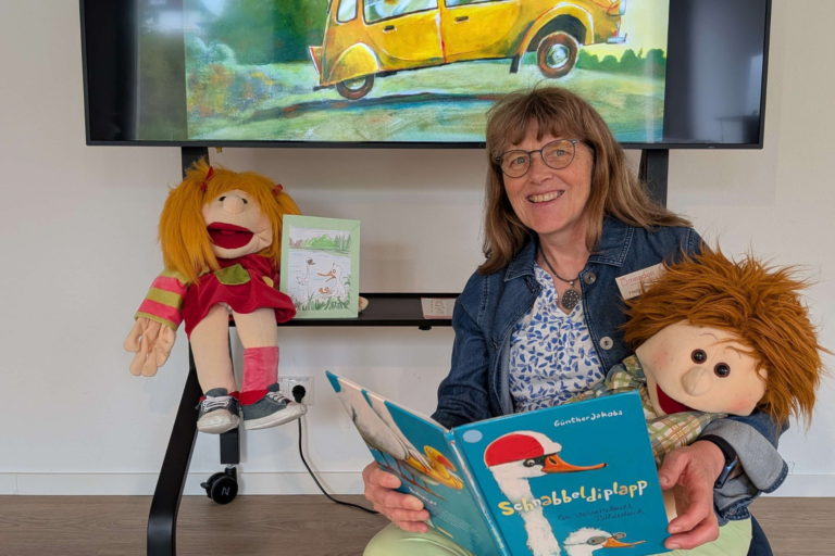 Foto: Dorte-Hilleke-Stadtbibliothek Eine lächelnde Frau sitzt mit zwei Handpuppen vor einem großen Bildschirm und hält das Bilderbuch „Schnabbeldiplapp“ geöffnet in den Händen. Auf dem Bildschirm ist eine farbenfrohe Illustration aus dem Buch mit zwei Gänsen in einem gelben Auto zu sehen.