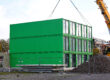 Ein dreistöckiges Gebäude aus grünen Containermodulen steht auf einer Baustelle. Ein großer gelber Autokran hebt ein weiteres Modul mit Stahlseilen an. Rechts sind eine Arbeitsbühne und ein großer Schutthaufen zu sehen. Bauzäune sichern das Gelände ab.