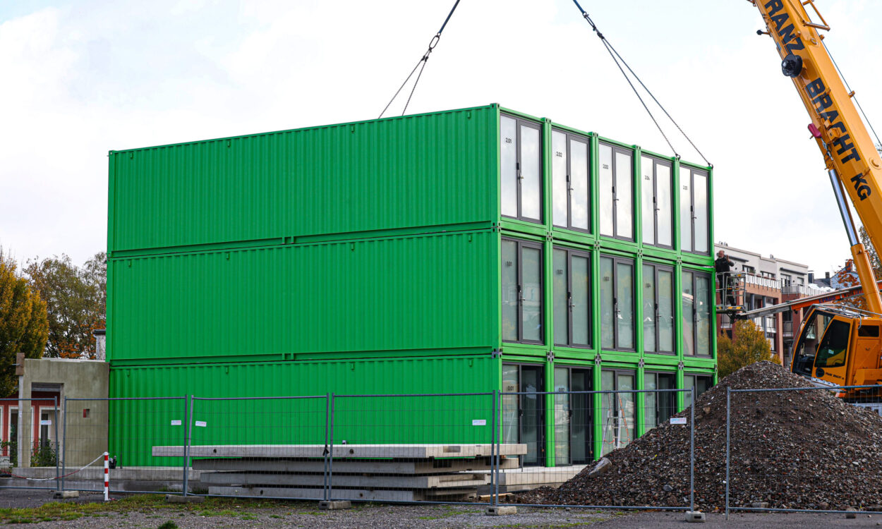 Ein dreistöckiges Gebäude aus grünen Containermodulen steht auf einer Baustelle. Ein großer gelber Autokran hebt ein weiteres Modul mit Stahlseilen an. Rechts sind eine Arbeitsbühne und ein großer Schutthaufen zu sehen. Bauzäune sichern das Gelände ab.