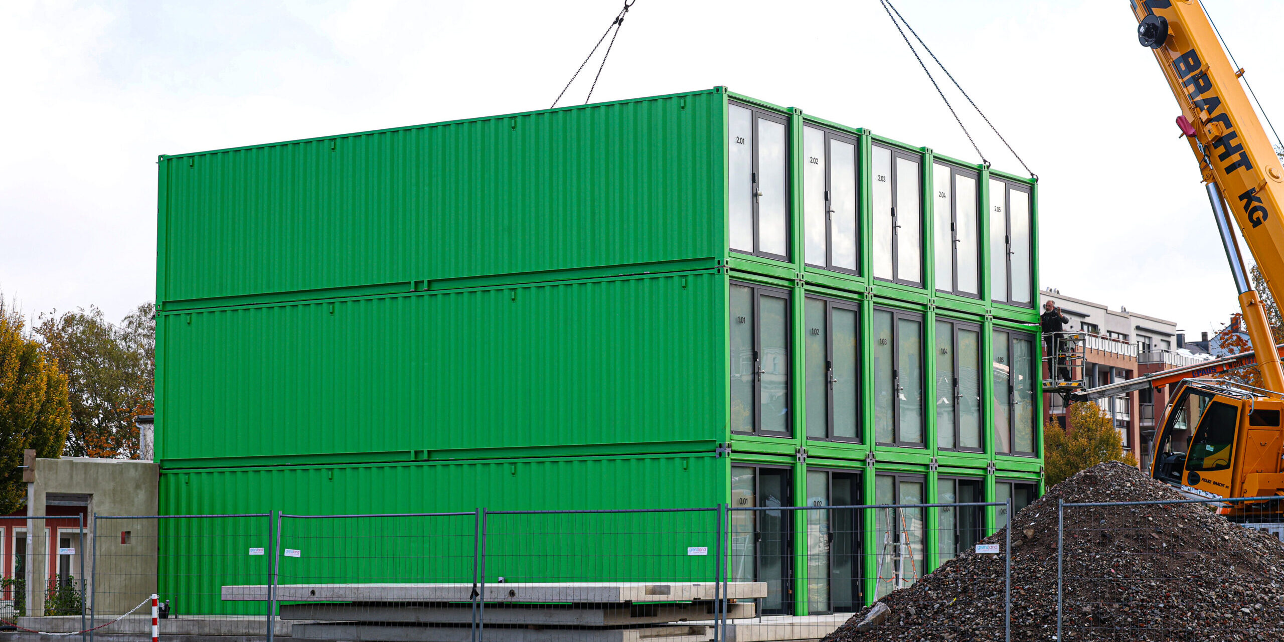Ein dreistöckiges Gebäude aus grünen Containermodulen steht auf einer Baustelle. Ein großer gelber Autokran hebt ein weiteres Modul mit Stahlseilen an. Rechts sind eine Arbeitsbühne und ein großer Schutthaufen zu sehen. Bauzäune sichern das Gelände ab.
