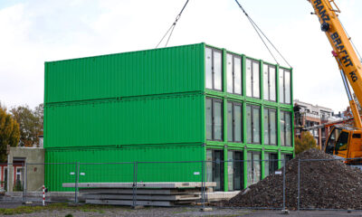 Ein dreistöckiges Gebäude aus grünen Containermodulen steht auf einer Baustelle. Ein großer gelber Autokran hebt ein weiteres Modul mit Stahlseilen an. Rechts sind eine Arbeitsbühne und ein großer Schutthaufen zu sehen. Bauzäune sichern das Gelände ab.