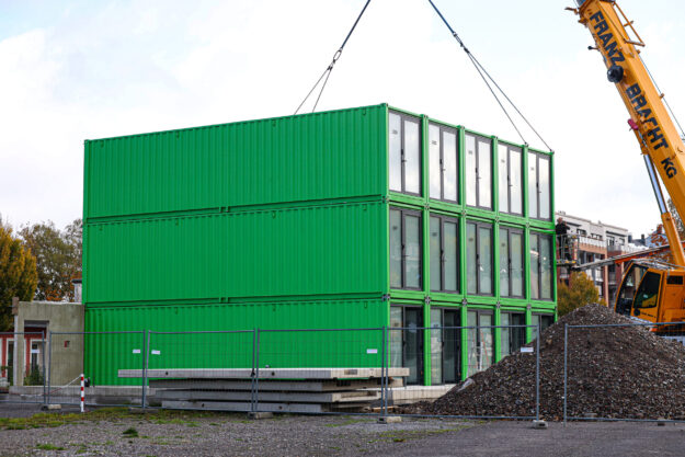 Foto: Dennis Echtermann Ein dreistöckiges Gebäude aus grünen Containermodulen steht auf einer Baustelle. Ein großer gelber Autokran hebt ein weiteres Modul mit Stahlseilen an. Rechts sind eine Arbeitsbühne und ein großer Schutthaufen zu sehen. Bauzäune sichern das Gelände ab.
