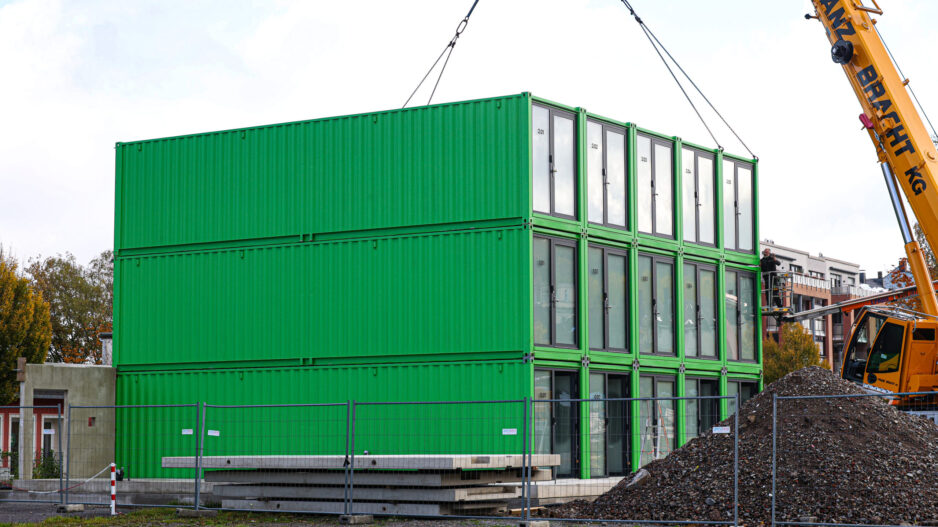 Ein dreistöckiges Gebäude aus grünen Containermodulen steht auf einer Baustelle. Ein großer gelber Autokran hebt ein weiteres Modul mit Stahlseilen an. Rechts sind eine Arbeitsbühne und ein großer Schutthaufen zu sehen. Bauzäune sichern das Gelände ab.