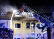 Nächtlicher Feuerwehreinsatz an einem Wohnhaus in Iserlohn. Aus dem Dach steigt Rauch auf, während Einsatzkräfte der Feuerwehr mit einer ausgefahrenen Drehleiter den Brand bekämpfen. Blaue Einsatzlichter beleuchten das Gebäude, vor dem ein Feuerwehrmann Anweisungen gibt.
