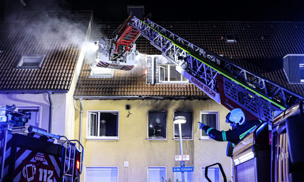 Nächtlicher Feuerwehreinsatz an einem Wohnhaus in Iserlohn. Aus dem Dach steigt Rauch auf, während Einsatzkräfte der Feuerwehr mit einer ausgefahrenen Drehleiter den Brand bekämpfen. Blaue Einsatzlichter beleuchten das Gebäude, vor dem ein Feuerwehrmann Anweisungen gibt.