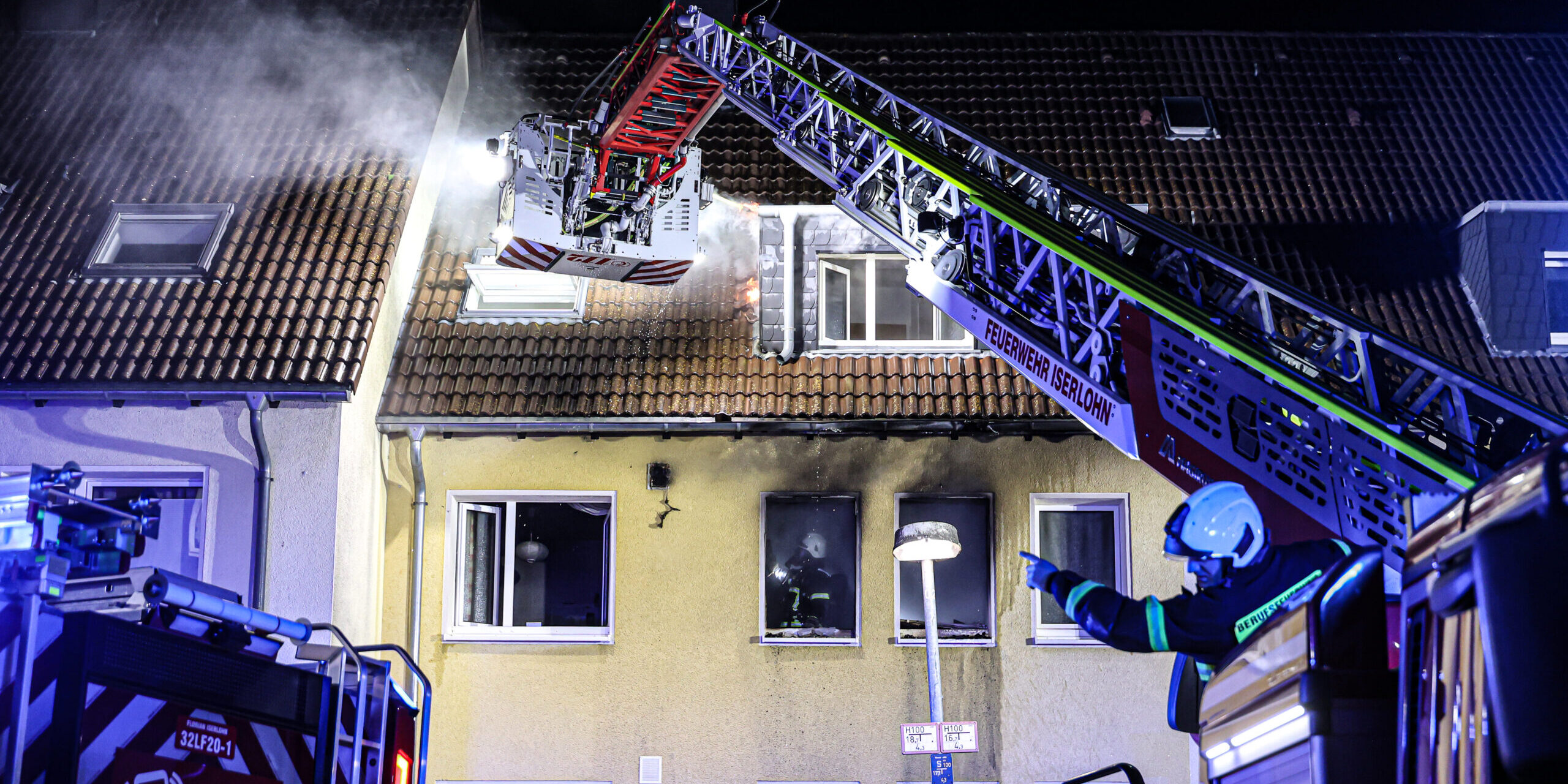 Nächtlicher Feuerwehreinsatz an einem Wohnhaus in Iserlohn. Aus dem Dach steigt Rauch auf, während Einsatzkräfte der Feuerwehr mit einer ausgefahrenen Drehleiter den Brand bekämpfen. Blaue Einsatzlichter beleuchten das Gebäude, vor dem ein Feuerwehrmann Anweisungen gibt.