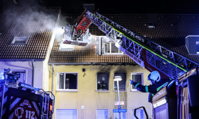 Nächtlicher Feuerwehreinsatz an einem Wohnhaus in Iserlohn. Aus dem Dach steigt Rauch auf, während Einsatzkräfte der Feuerwehr mit einer ausgefahrenen Drehleiter den Brand bekämpfen. Blaue Einsatzlichter beleuchten das Gebäude, vor dem ein Feuerwehrmann Anweisungen gibt.