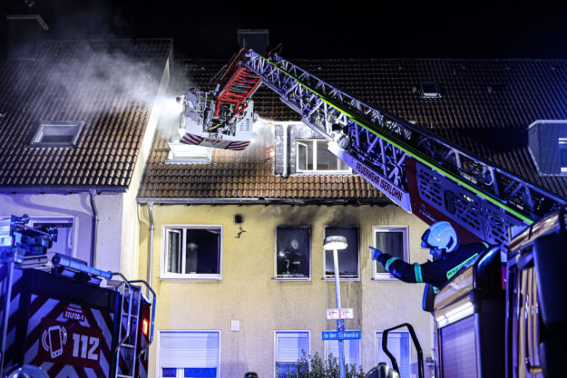 Nächtlicher Feuerwehreinsatz an einem Wohnhaus in Iserlohn. Aus dem Dach steigt Rauch auf, während Einsatzkräfte der Feuerwehr mit einer ausgefahrenen Drehleiter den Brand bekämpfen. Blaue Einsatzlichter beleuchten das Gebäude, vor dem ein Feuerwehrmann Anweisungen gibt.