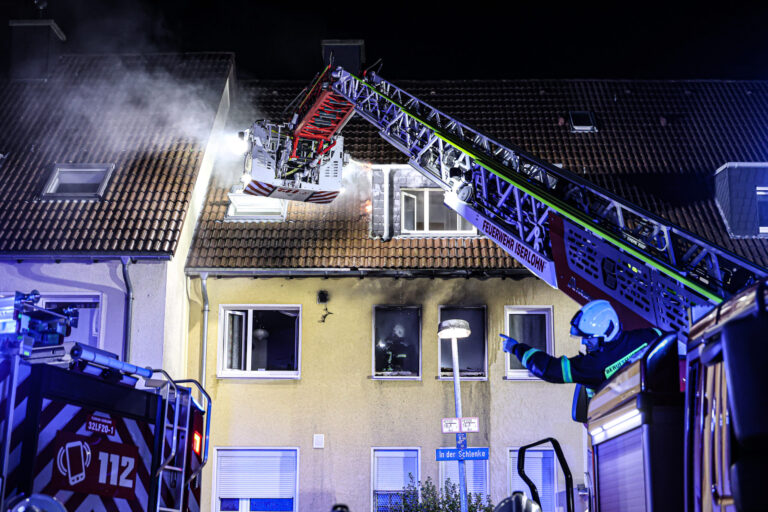 Foto: Dennis Echtermann Nächtlicher Feuerwehreinsatz an einem Wohnhaus in Iserlohn. Aus dem Dach steigt Rauch auf, während Einsatzkräfte der Feuerwehr mit einer ausgefahrenen Drehleiter den Brand bekämpfen. Blaue Einsatzlichter beleuchten das Gebäude, vor dem ein Feuerwehrmann Anweisungen gibt.