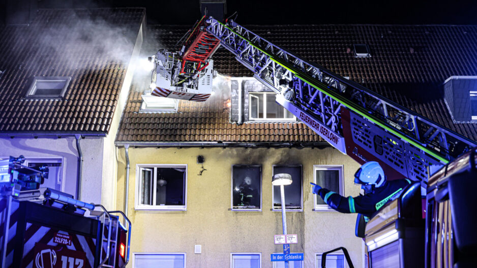 Nächtlicher Feuerwehreinsatz an einem Wohnhaus in Iserlohn. Aus dem Dach steigt Rauch auf, während Einsatzkräfte der Feuerwehr mit einer ausgefahrenen Drehleiter den Brand bekämpfen. Blaue Einsatzlichter beleuchten das Gebäude, vor dem ein Feuerwehrmann Anweisungen gibt.