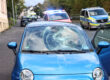 Ein blauer Kleinwagen steht mit stark beschädigter Windschutzscheibe und eingedellter Motorhaube am Straßenrand. Die Beifahrertür ist geöffnet. Im Hintergrund stehen ein Rettungswagen mit eingeschaltetem Blaulicht, ein Polizeifahrzeug und ein weiterer Rettungswagen.