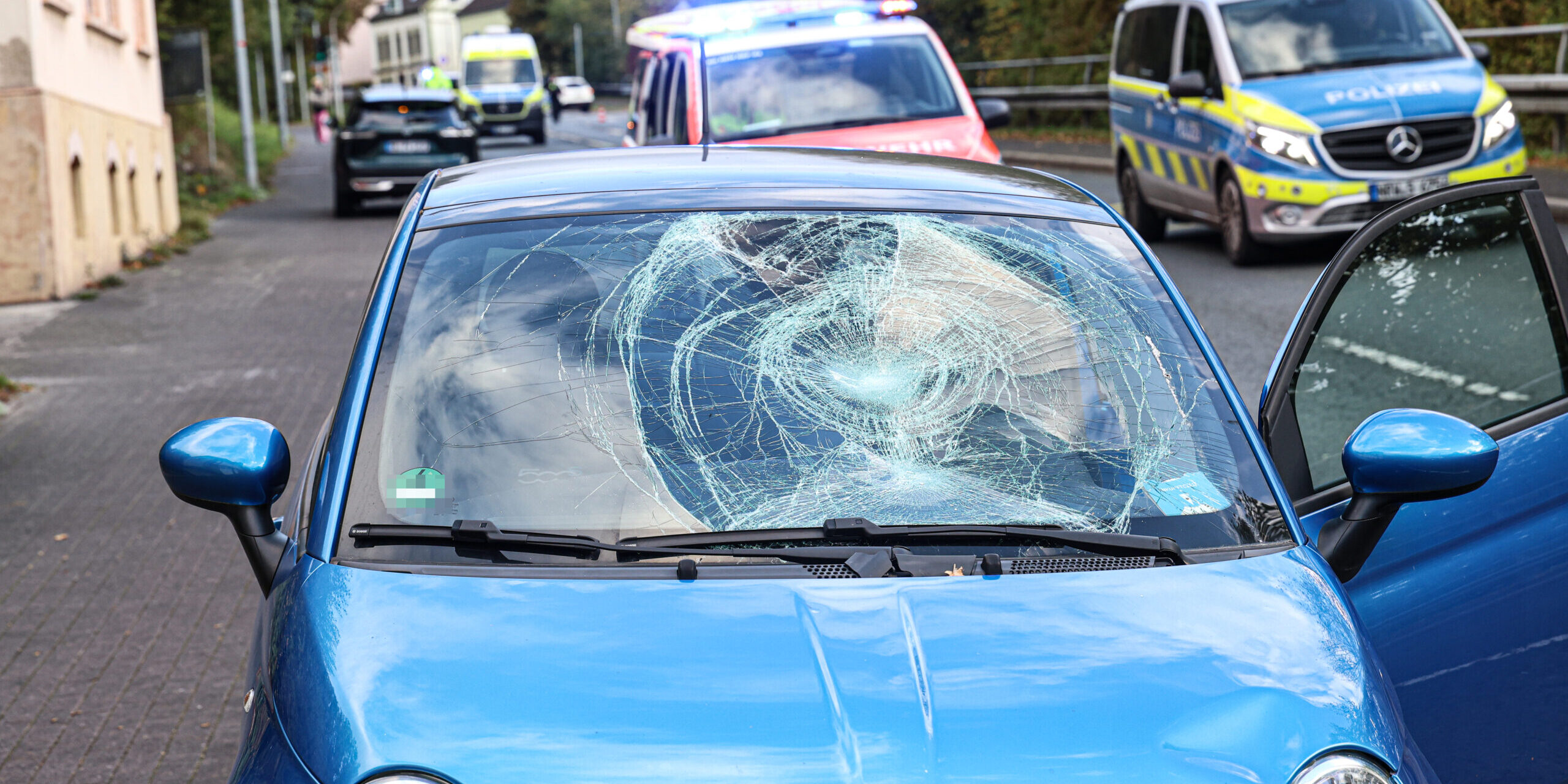 Ein blauer Kleinwagen steht mit stark beschädigter Windschutzscheibe und eingedellter Motorhaube am Straßenrand. Die Beifahrertür ist geöffnet. Im Hintergrund stehen ein Rettungswagen mit eingeschaltetem Blaulicht, ein Polizeifahrzeug und ein weiterer Rettungswagen.