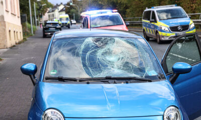 Ein blauer Kleinwagen steht mit stark beschädigter Windschutzscheibe und eingedellter Motorhaube am Straßenrand. Die Beifahrertür ist geöffnet. Im Hintergrund stehen ein Rettungswagen mit eingeschaltetem Blaulicht, ein Polizeifahrzeug und ein weiterer Rettungswagen.