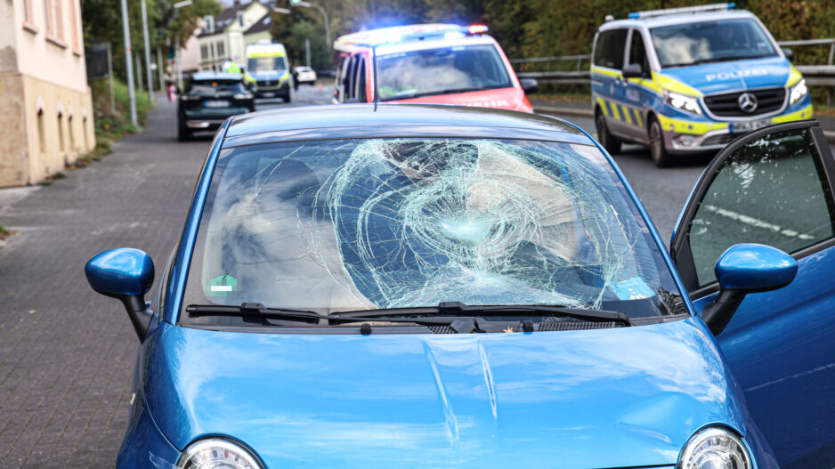 Ein blauer Kleinwagen steht mit stark beschädigter Windschutzscheibe und eingedellter Motorhaube am Straßenrand. Die Beifahrertür ist geöffnet. Im Hintergrund stehen ein Rettungswagen mit eingeschaltetem Blaulicht, ein Polizeifahrzeug und ein weiterer Rettungswagen.