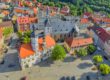 Luftaufnahme der historischen Innenstadt von Eisenberg in Thüringen. Im Mittelpunkt stehen das Rathaus mit seinem markanten Uhrturm, eine große Kirche und ein weitläufiger, gepflasterter Marktplatz mit Bäumen. Rundherum befinden sich historische Gebäude mit überwiegend roten Ziegeldächern.
