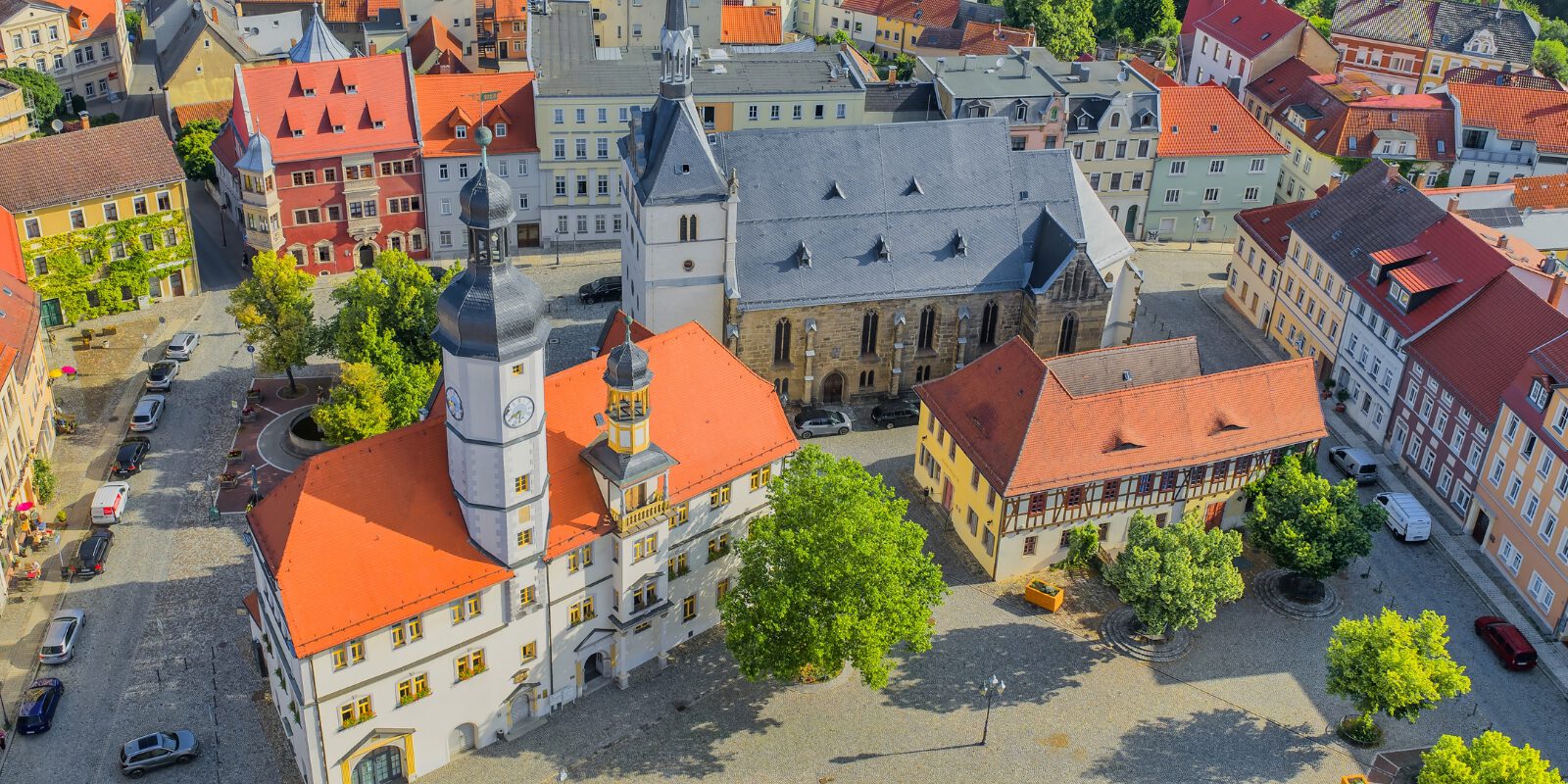 Luftaufnahme der historischen Innenstadt von Eisenberg in Thüringen. Im Mittelpunkt stehen das Rathaus mit seinem markanten Uhrturm, eine große Kirche und ein weitläufiger, gepflasterter Marktplatz mit Bäumen. Rundherum befinden sich historische Gebäude mit überwiegend roten Ziegeldächern.