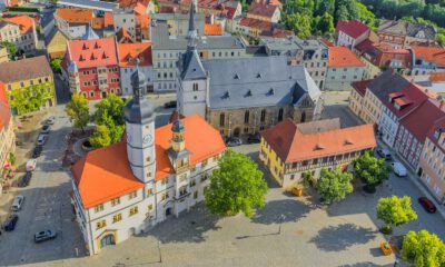 Luftaufnahme der historischen Innenstadt von Eisenberg in Thüringen. Im Mittelpunkt stehen das Rathaus mit seinem markanten Uhrturm, eine große Kirche und ein weitläufiger, gepflasterter Marktplatz mit Bäumen. Rundherum befinden sich historische Gebäude mit überwiegend roten Ziegeldächern.
