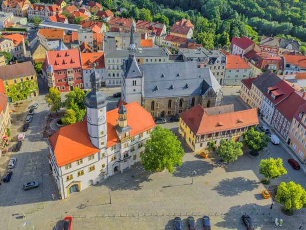 Luftaufnahme der historischen Innenstadt von Eisenberg in Thüringen. Im Mittelpunkt stehen das Rathaus mit seinem markanten Uhrturm, eine große Kirche und ein weitläufiger, gepflasterter Marktplatz mit Bäumen. Rundherum befinden sich historische Gebäude mit überwiegend roten Ziegeldächern.