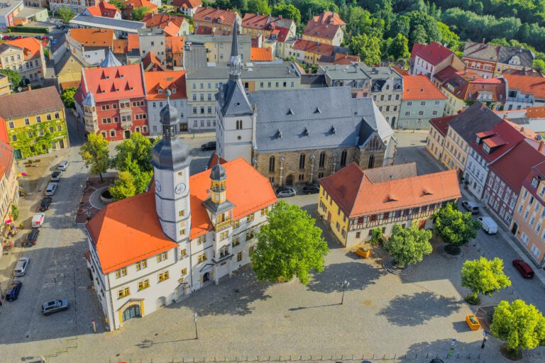 Luftaufnahme der historischen Innenstadt von Eisenberg in Thüringen. Im Mittelpunkt stehen das Rathaus mit seinem markanten Uhrturm, eine große Kirche und ein weitläufiger, gepflasterter Marktplatz mit Bäumen. Rundherum befinden sich historische Gebäude mit überwiegend roten Ziegeldächern.