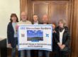 Fünf Personen stehen vor einer Holztür und präsentieren ein großes Protestplakat gegen Windkraftanlagen in Sümmern und auf dem Gaxberg. Das Plakat zeigt zwei Windräder und trägt die Aufschrift „592 Menschen fordern Windrad-Stopp in Sümmern und auf’m Gaxberg!“.