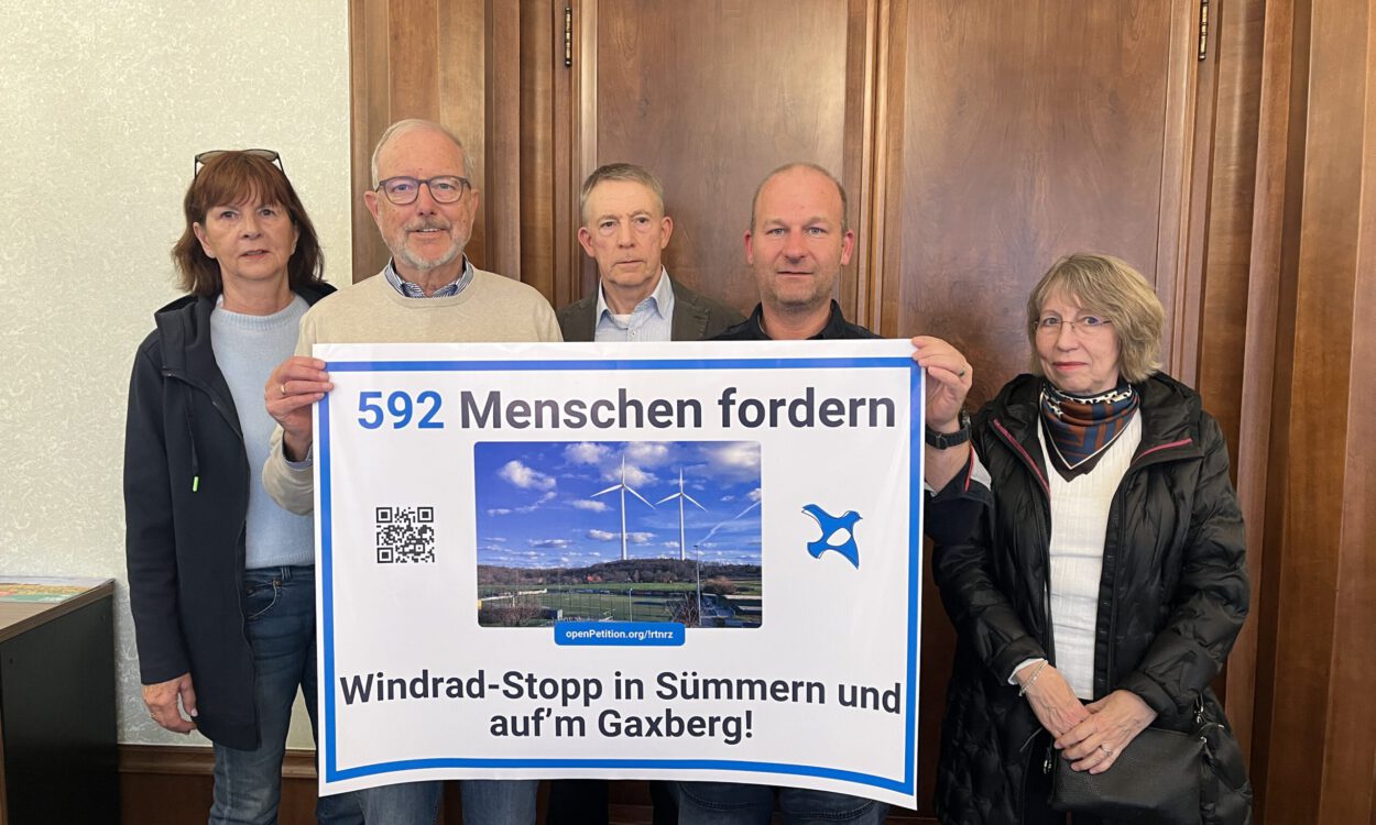 Fünf Personen stehen vor einer Holztür und präsentieren ein großes Protestplakat gegen Windkraftanlagen in Sümmern und auf dem Gaxberg. Das Plakat zeigt zwei Windräder und trägt die Aufschrift „592 Menschen fordern Windrad-Stopp in Sümmern und auf’m Gaxberg!“.