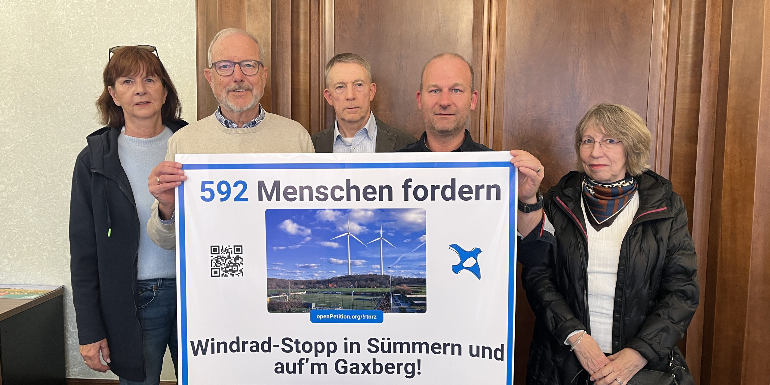 Fünf Personen stehen vor einer Holztür und präsentieren ein großes Protestplakat gegen Windkraftanlagen in Sümmern und auf dem Gaxberg. Das Plakat zeigt zwei Windräder und trägt die Aufschrift „592 Menschen fordern Windrad-Stopp in Sümmern und auf’m Gaxberg!“.
