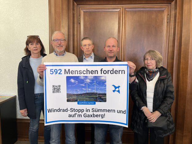 Foto: Sabrina Fehring Fünf Personen stehen vor einer Holztür und präsentieren ein großes Protestplakat gegen Windkraftanlagen in Sümmern und auf dem Gaxberg. Das Plakat zeigt zwei Windräder und trägt die Aufschrift „592 Menschen fordern Windrad-Stopp in Sümmern und auf’m Gaxberg!“.