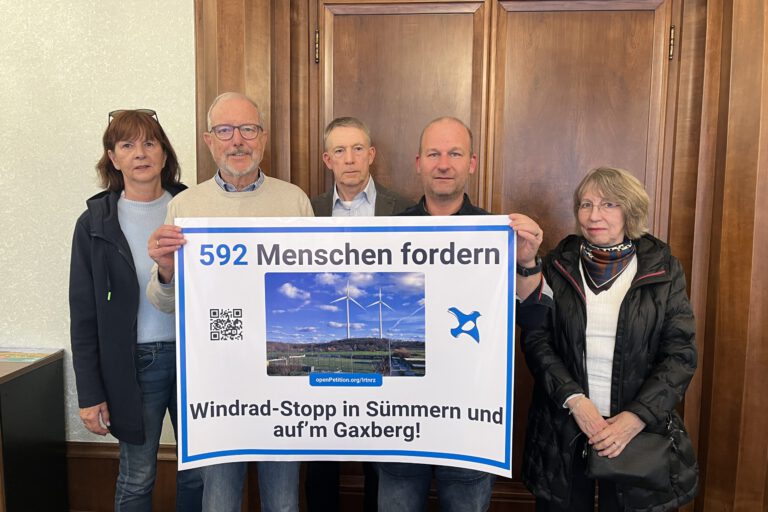 Fünf Personen stehen vor einer Holztür und präsentieren ein großes Protestplakat gegen Windkraftanlagen in Sümmern und auf dem Gaxberg. Das Plakat zeigt zwei Windräder und trägt die Aufschrift „592 Menschen fordern Windrad-Stopp in Sümmern und auf’m Gaxberg!“.