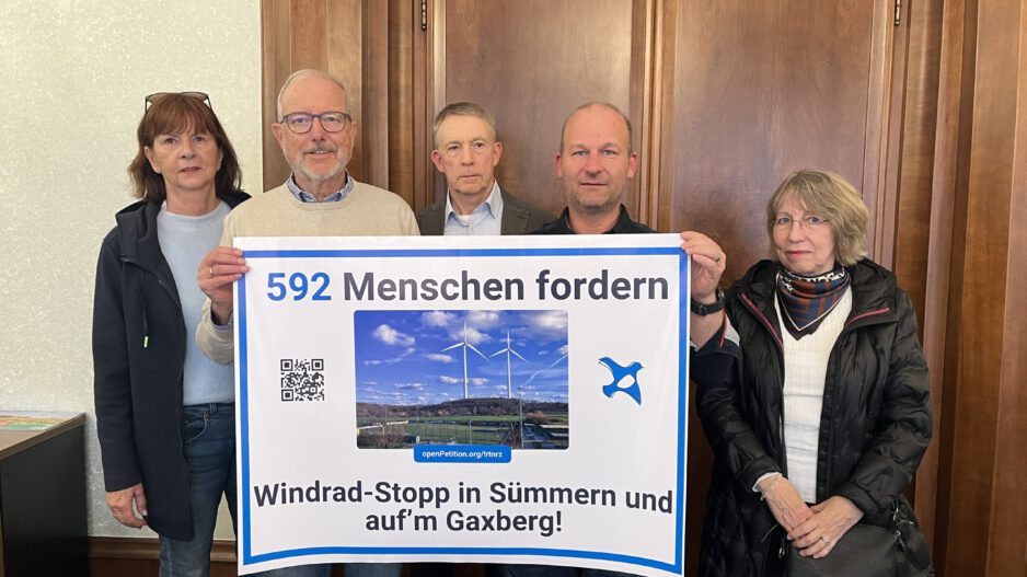 Fünf Personen stehen vor einer Holztür und präsentieren ein großes Protestplakat gegen Windkraftanlagen in Sümmern und auf dem Gaxberg. Das Plakat zeigt zwei Windräder und trägt die Aufschrift „592 Menschen fordern Windrad-Stopp in Sümmern und auf’m Gaxberg!“.