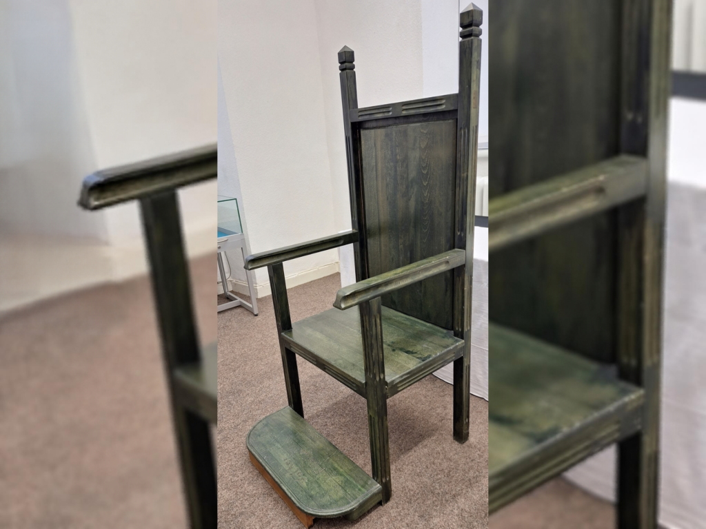 Ein hoher, dunkel gebeizter Holzstuhl mit Armlehnen, markanter hoher Rückenlehne und vier kantigen Pfosten steht in einem Ausstellungsraum. Vor dem Sitz befindet sich eine niedrige, abgerundete Fußstütze.