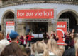 Vor einem historischen Gebäude treten mehrere Kinder und Jugendliche in roten und dunkelroten Oberteilen auf. Zahlreiche Zuschauerinnen und Zuschauer verfolgen die Darbietung. Über der Bühne hängt ein großes rotes Banner mit der Aufschrift „tor zur vielfalt“.