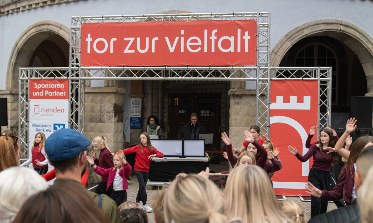 Vor einem historischen Gebäude treten mehrere Kinder und Jugendliche in roten und dunkelroten Oberteilen auf. Zahlreiche Zuschauerinnen und Zuschauer verfolgen die Darbietung. Über der Bühne hängt ein großes rotes Banner mit der Aufschrift „tor zur vielfalt“.
