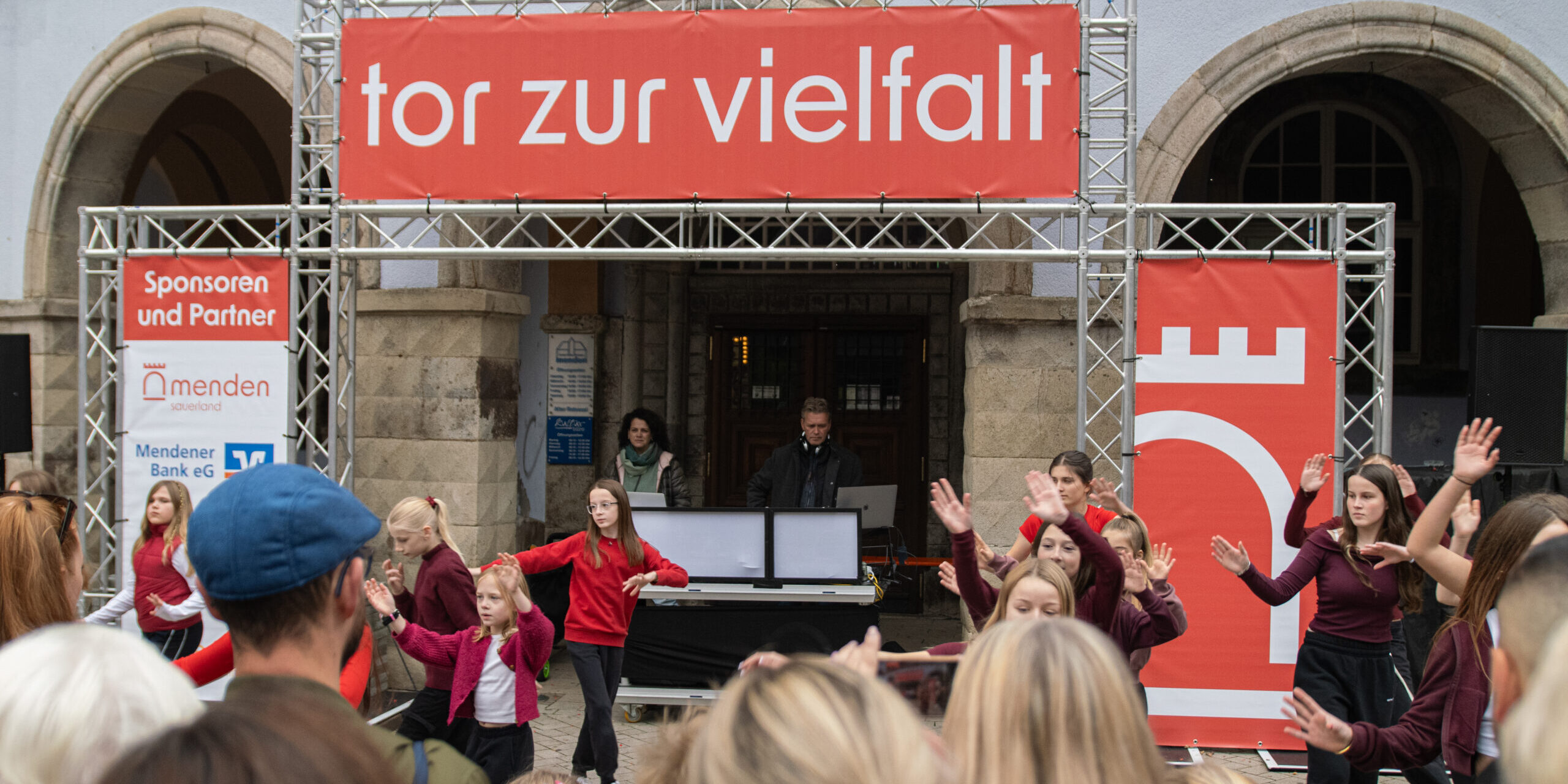 Vor einem historischen Gebäude treten mehrere Kinder und Jugendliche in roten und dunkelroten Oberteilen auf. Zahlreiche Zuschauerinnen und Zuschauer verfolgen die Darbietung. Über der Bühne hängt ein großes rotes Banner mit der Aufschrift „tor zur vielfalt“.