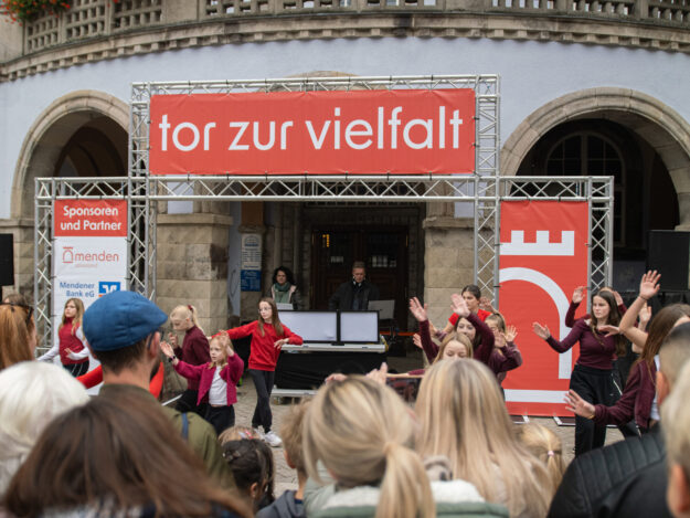 Foto: Jannik Zirbel/Stadtmarketing Menden Vor einem historischen Gebäude treten mehrere Kinder und Jugendliche in roten und dunkelroten Oberteilen auf. Zahlreiche Zuschauerinnen und Zuschauer verfolgen die Darbietung. Über der Bühne hängt ein großes rotes Banner mit der Aufschrift „tor zur vielfalt“.